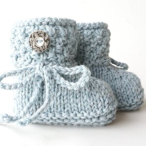 May include: A pair of light blue knitted baby booties. The booties have a textured stitch pattern, a decorative button, and a tie closure. The soft blue color and handmade appearance suggest a cozy, handcrafted item, perfect for a newborn.