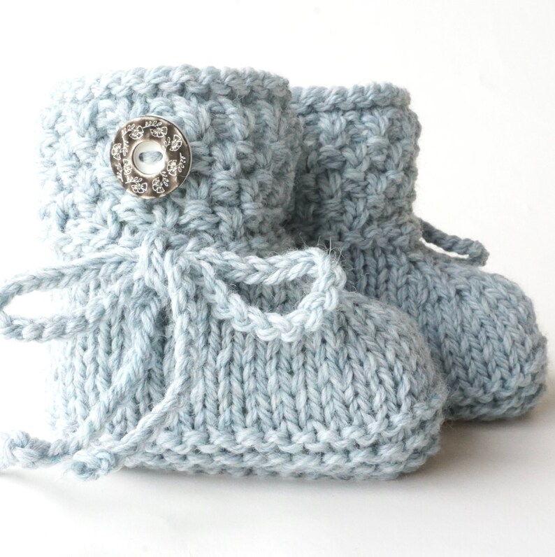 May include: A pair of light blue knitted baby booties. The booties have a textured stitch pattern, a decorative button, and a tie closure. The soft blue color and handmade appearance suggest a cozy, handcrafted item, perfect for a newborn.