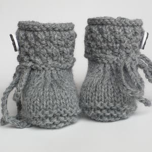 May include: A pair of gray knitted baby booties. The booties have a cuff with a textured pattern and a small button detail. Each bootie has a drawstring. The booties are made of a soft, gray yarn.