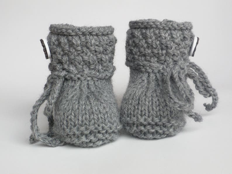 May include: A pair of gray knitted baby booties. The booties have a cuff with a textured pattern and a small button detail. Each bootie has a drawstring. The booties are made of a soft, gray yarn.
