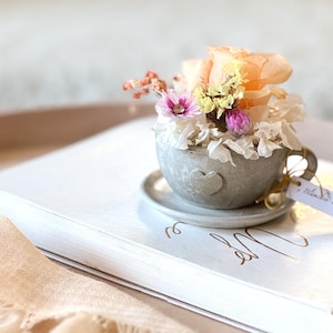 Könnte beinhalten: Eine kleine, graue Teetasse gefüllt mit getrockneten Blumen in Pfirsich-, Rosa- und Gelbtönen, die auf einer Untertasse ruht. Die Tasse steht auf einem weißen Buch, mit einem Etikett mit der Aufschrift "Blonde Koffie".