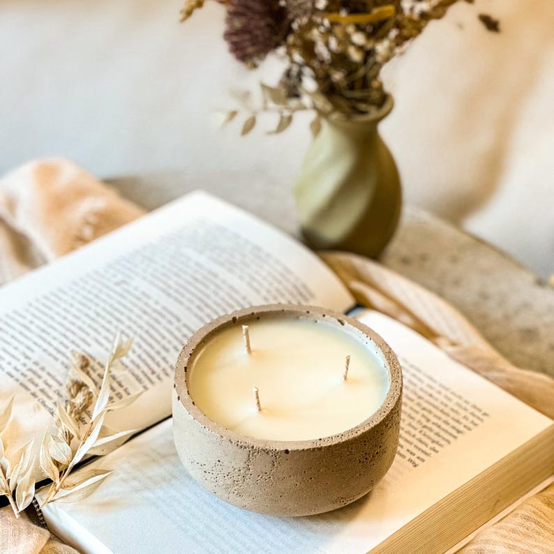 May include: A beige concrete candle holder with three wicks and a white candle. The candle is sitting on a book with a dried flower arrangement in the background.