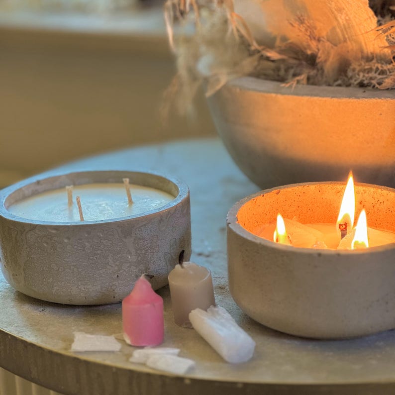 May include: Three concrete candle holders with lit candles. The candles are in various sizes and shapes. The candle holders are a light gray color and have a rough texture.