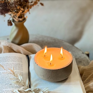 May include: A three-wick concrete candle with a warm, orange glow. The candle is sitting on an open book.