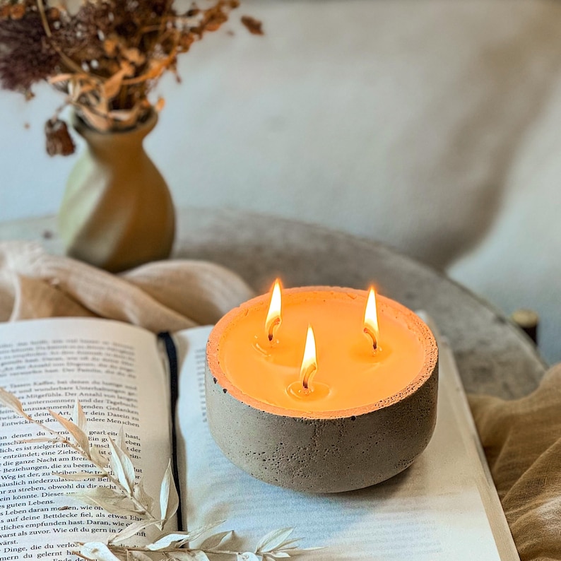 May include: A three-wick concrete candle with a warm, orange glow. The candle is sitting on an open book.