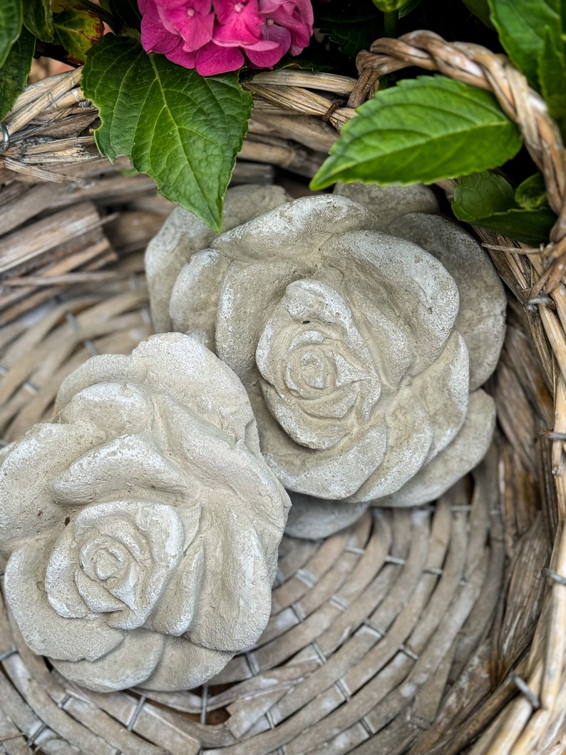Op de afbeelding: Twee grijze betonnen rozen sculpturen in een rieten mand. De rozen zijn gedetailleerd en realistisch.