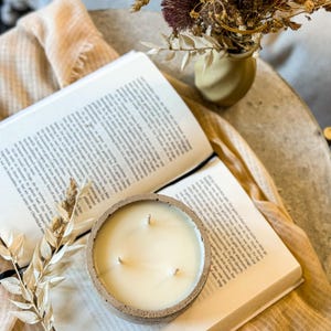 May include: A white concrete candle with three wicks sits on top of an open book. The candle is surrounded by dried flowers and a beige fabric.