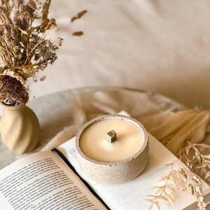 Op de afbeelding: Een beige betonnen kaarsenhouder met een witte kaars en een houten lont. De kaarsenhouder staat op een boek met gedroogde bloemen op de achtergrond.