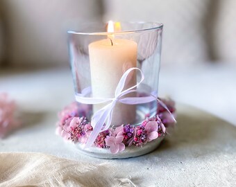 May include: A white candle in a clear glass holder with a white ribbon and a wreath of pink and purple flowers around the base.