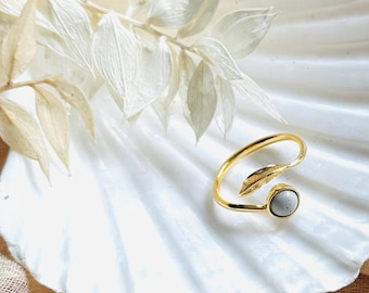 May include: A gold-toned open ring featuring a polished metal leaf design on one end and a small, smooth, circular grey stone setting on the other end. The ring is crafted with a slender, curved band that wraps around the finger.