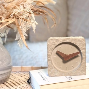 Könnte beinhalten: Eine quadratische, beige Stein-Uhr mit hellbeigem Zifferblatt und Holzzeigern. Die Uhr steht auf einem Stapel Bücher neben einer Vase mit Trockenblumen. Das Design der Uhr ist minimalistisch und modern.