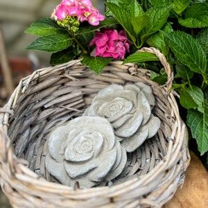 Op de afbeelding: Twee grijze betonnen rozenvormige tuinversieringen in een rieten mand met roze bloemen.