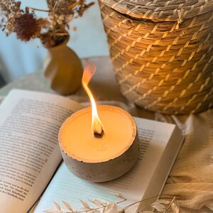 Op de afbeelding: Een betonnen kaarsenhouder met een brandende kaars erin. De kaars is lichtgeel en de houder is lichtgrijs. De kaars staat op een boek.