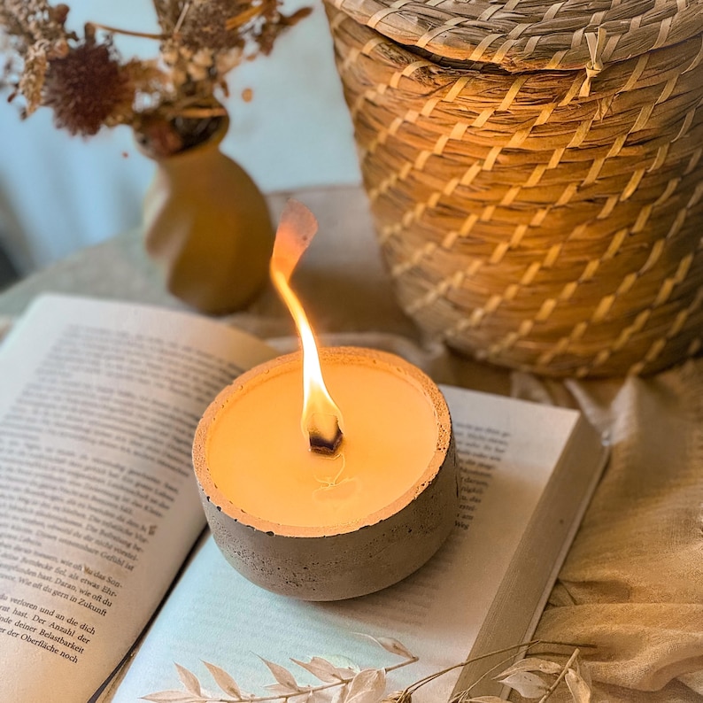 Op de afbeelding: Een betonnen kaarsenhouder met een brandende kaars erin. De kaars is lichtgeel en de houder is lichtgrijs. De kaars staat op een boek.