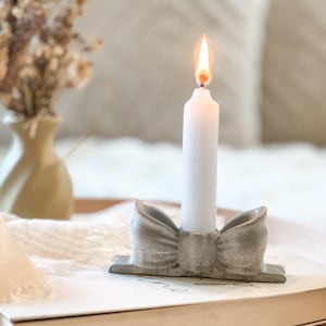 May include: A gray ceramic candle holder shaped like a bow, holding a single white taper candle with a lit flame. The candle holder has a smooth, glossy finish and sits on a flat surface, providing a stable base for the upright candle.