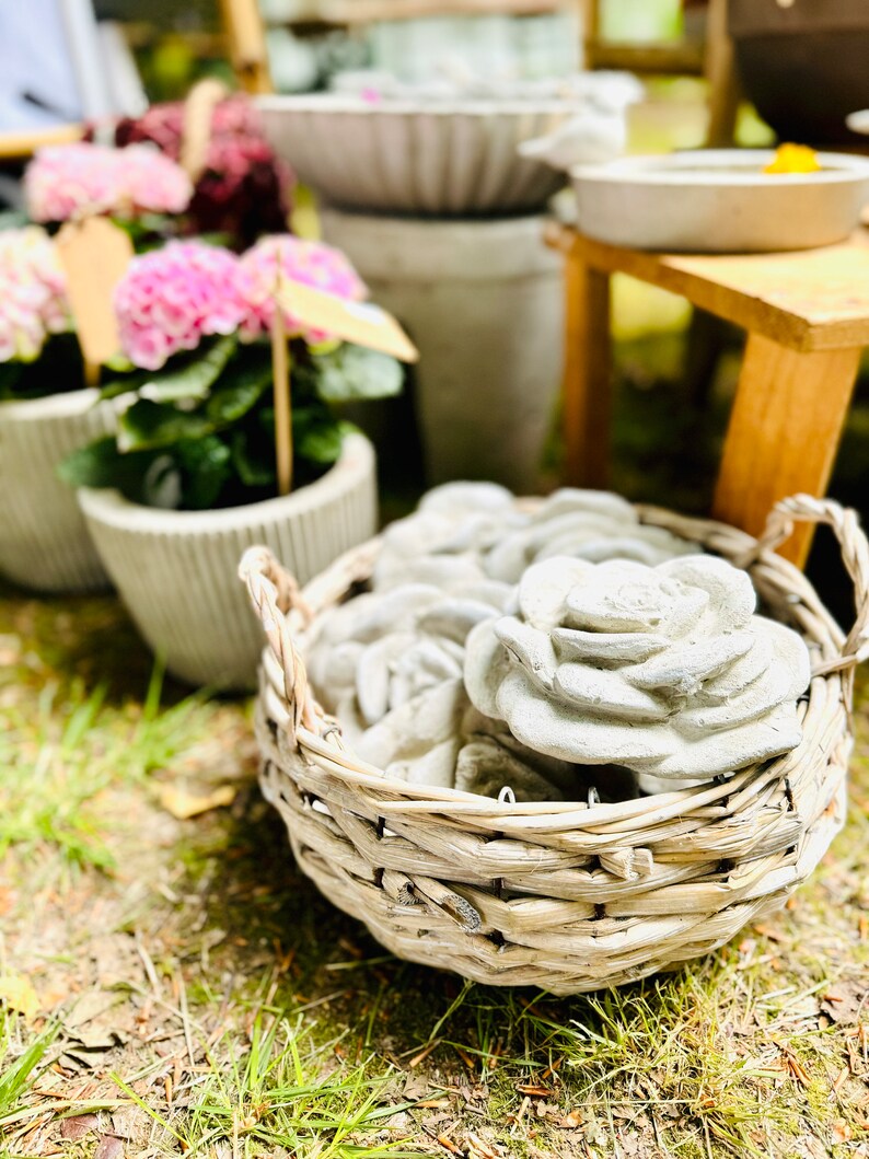 Op de afbeelding: Een rieten mand gevuld met grijze betonnen rozen en bloemenvormen. De mand staat op een bed van groen gras.