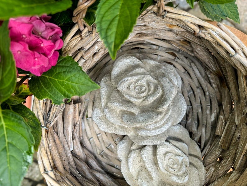 Op de afbeelding: Twee grijze betonnen rozen in een geweven rieten mand. De rozen zijn gedetailleerd met realistische bloemblaadjes en liggen in de mand.