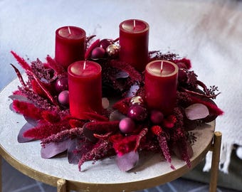 May include: A circular holiday wreath featuring four deep red pillar candles arranged among dark red dried grasses, feathery plumes, and eucalyptus leaves. The arrangement is accented with small, shiny, and matte burgundy and gold spherical ornaments.