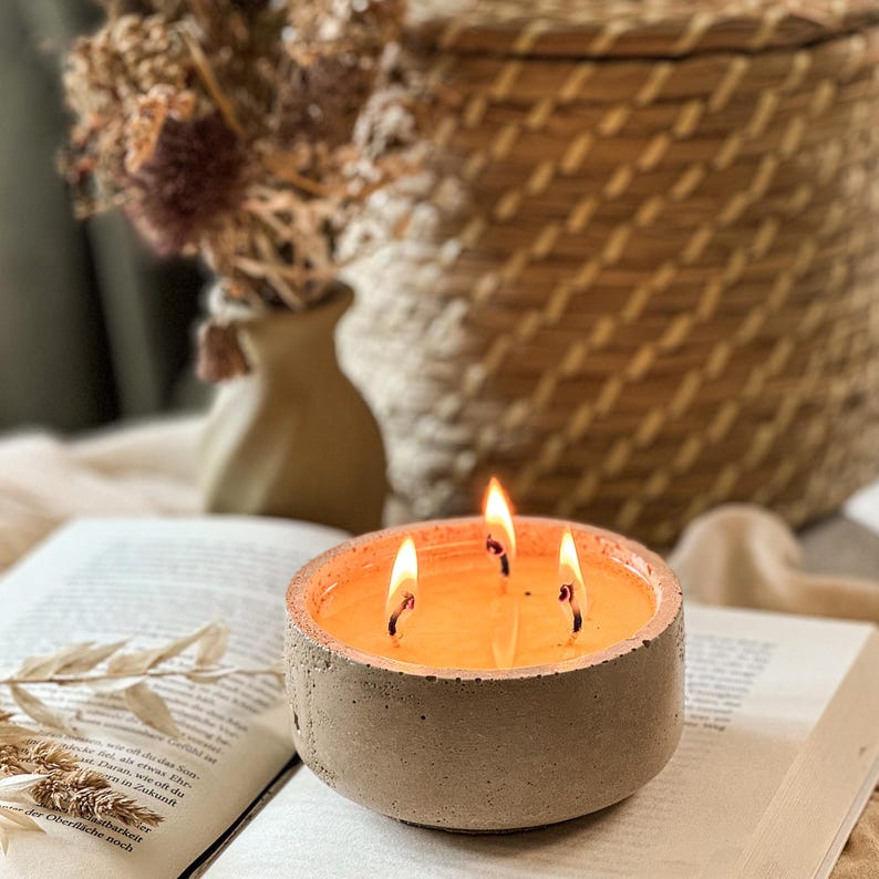 May include: A concrete candle holder with three lit wicks. The candle is a light orange color and is sitting on a book.