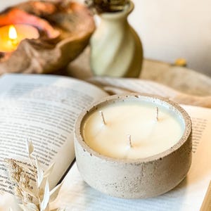 May include: A three-wick concrete candle with a beige exterior and a white wax interior. The candle is sitting on a book.