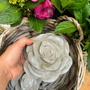 Op de afbeelding: Twee grijze betonnen rozen sculpturen in een rieten mand met roze hortensia's.