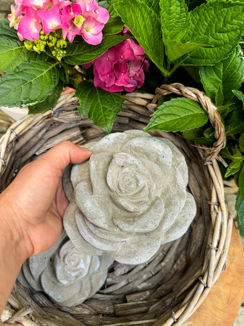 Op de afbeelding: Twee grijze betonnen rozen sculpturen in een rieten mand met roze hortensia's.