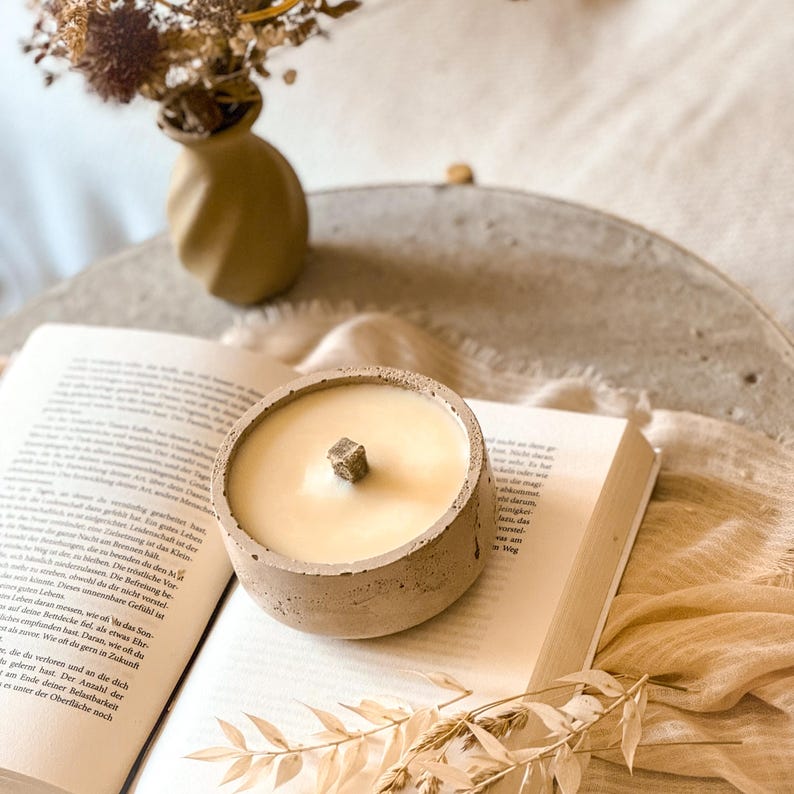 Op de afbeelding: Een beige betonnen kaarsenhouder met een beige kaars erin. De kaars heeft een houten lont. De kaarsenhouder staat op een open boek.