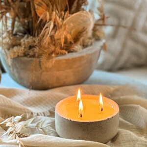 May include: A concrete candle holder with three lit wicks and a yellow wax pool. The candle holder is sitting on a beige fabric surface.
