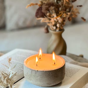 May include: A gray concrete candle holder with three lit wicks. The candle is filled with a light orange wax. The candle holder is sitting on a book.