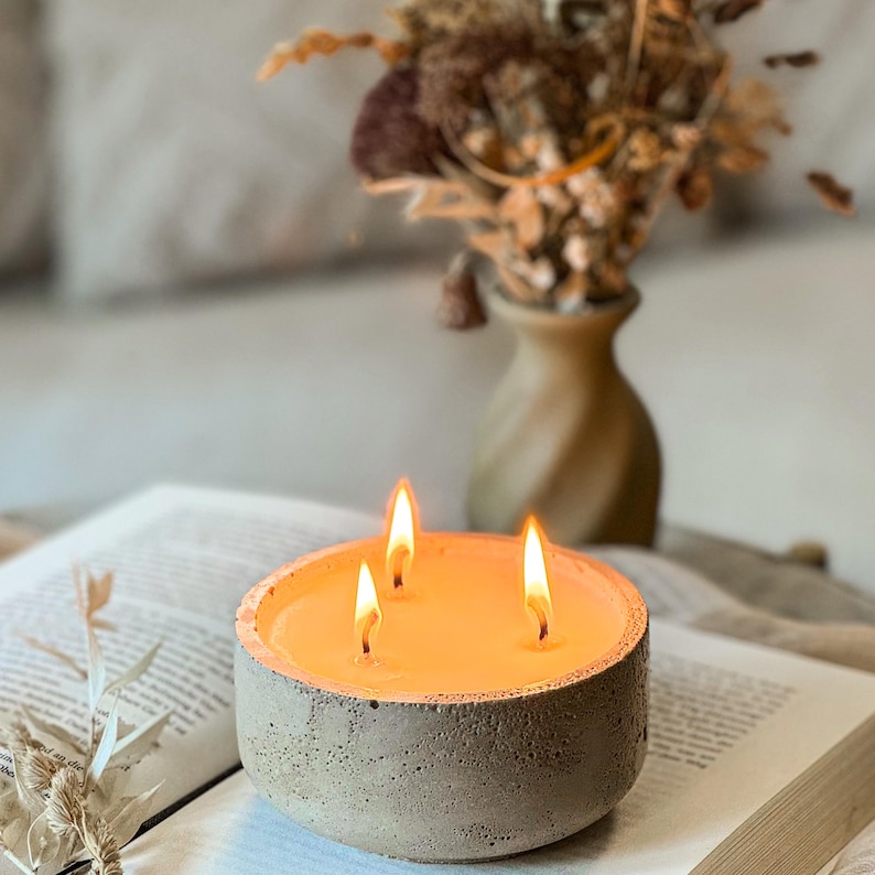 May include: A gray concrete candle holder with three lit wicks. The candle is filled with a light orange wax. The candle holder is sitting on a book.