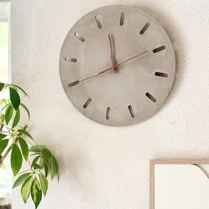 Könnte beinhalten: Eine runde, graue Betonuhr mit minimalistischem Design. Sie hat rechteckige Aussparungen am Rand als Stundenmarkierungen. Die Zeiger sind rotbraun. Die Uhr ist an einer strukturierten weißen Wand befestigt. Der Durchmesser beträgt ca. 30 cm.
