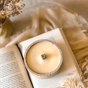 Op de afbeelding: Een betonnen kaarsenhouder met een beige, getextureerde afwerking. De kaars is wit en heeft een houten lont. De kaarsenhouder staat op een boek.