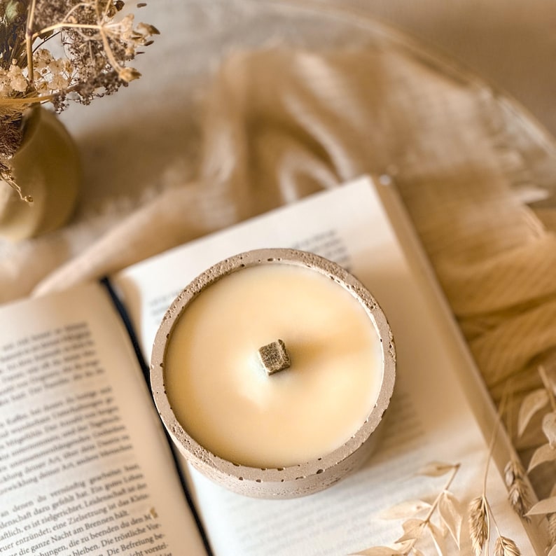 Op de afbeelding: Een betonnen kaarsenhouder met een beige, getextureerde afwerking. De kaars is wit en heeft een houten lont. De kaarsenhouder staat op een boek.