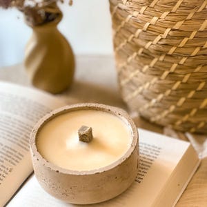 Op de afbeelding: Een beige betonnen kaarsenhouder met een lichtbeige kaars en een houten lont. De kaars staat op een boek.