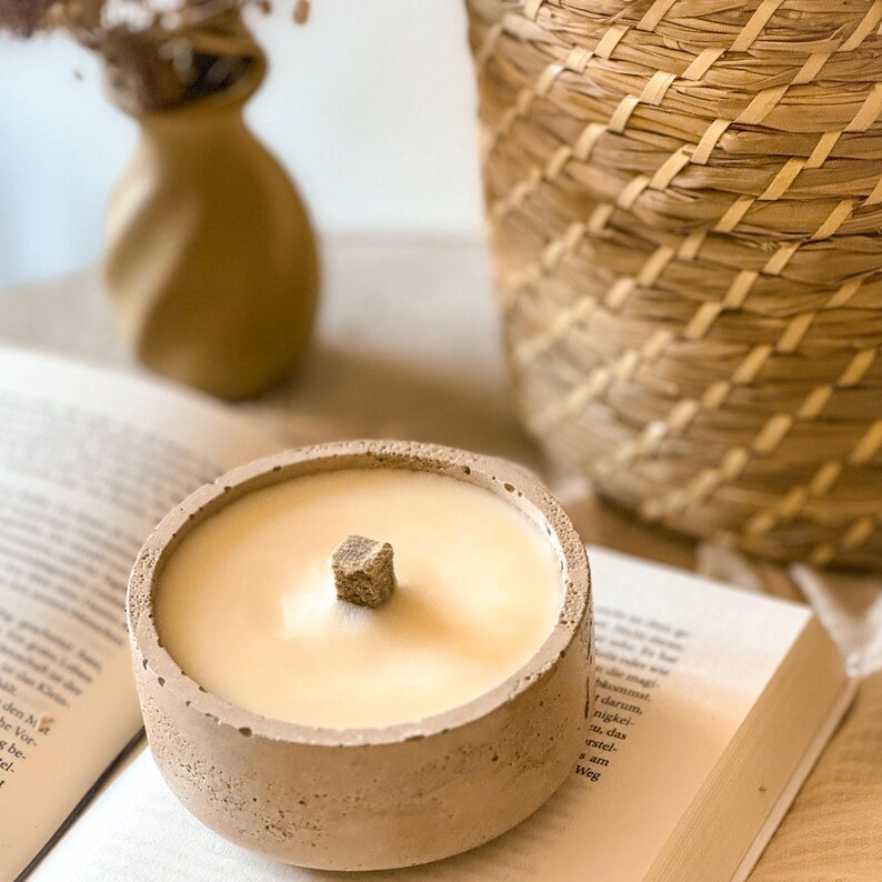 Op de afbeelding: Een beige betonnen kaarsenhouder met een lichtbeige kaars en een houten lont. De kaars staat op een boek.