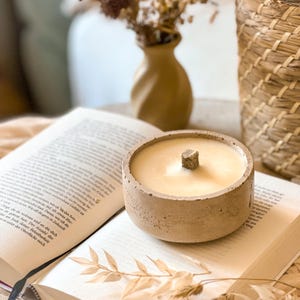 Op de afbeelding: Een beige betonnen kaarsenhouder met een beige wax kaars en een houten lont. De kaars staat op een open boek.