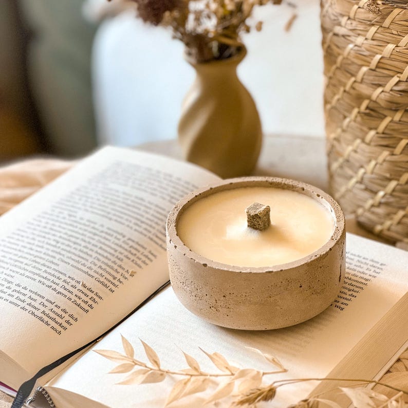 Op de afbeelding: Een beige betonnen kaarsenhouder met een beige wax kaars en een houten lont. De kaars staat op een open boek.