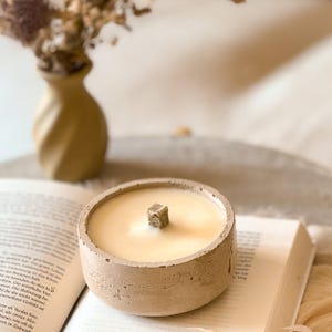 Op de afbeelding: Een beige betonnen kaarsenhouder met een beige kaars en een houten lont. De kaarsenhouder staat op een boek.