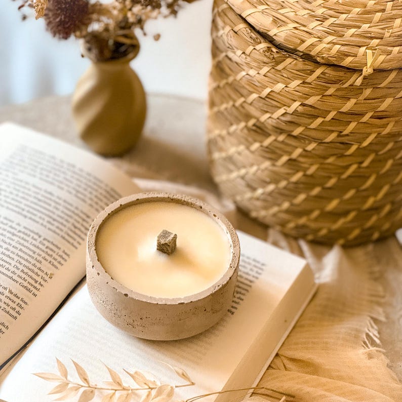 Op de afbeelding: Een beige betonnen kaarshouder met een lichtbeige kaars en een houten lont. De kaars staat op een open boek met een bruine geweven mand in de achtergrond.