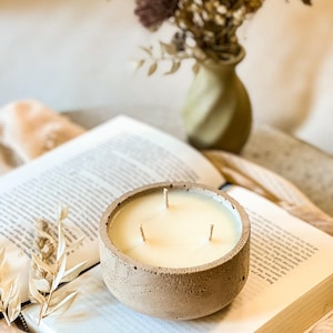 May include: A beige concrete candle holder with three wicks and a white candle. The candle is sitting on a book with a brown cover.