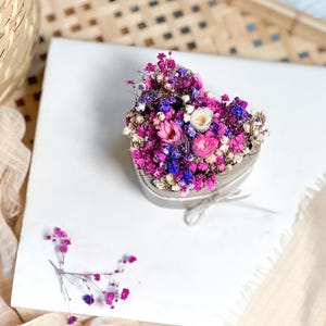 May include: A heart-shaped arrangement of dried flowers in shades of pink, purple, white, and blue. The floral display sits on a white surface, with a beige fabric and a woven basket in the background. The arrangement is tied with a beige ribbon.