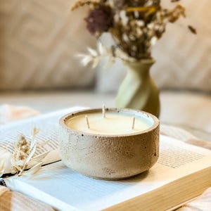 May include: A beige concrete candle holder with three wicks and a white candle. The candle is sitting on a book with a beige background.
