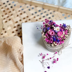 May include: A heart-shaped concrete container filled with dried flowers in shades of pink, purple, blue, and white. The container is tied with twine. A few loose flowers are scattered nearby. The arrangement sits on a white surface.