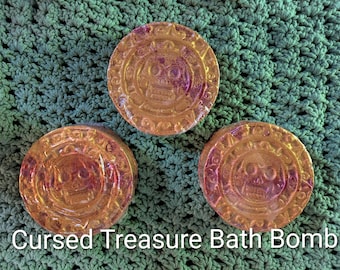 May include: Three round, orange-toned bath bombs with a metallic sheen, each featuring a raised skull design surrounded by an intricate border. The text Cursed Treasure Bath Bomb is printed in white at the bottom of the image.
