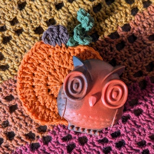 May include: A crocheted pumpkin with a brown stem and green leaves, sitting on a multi-colored crocheted blanket. A brown and orange owl figurine is next to the pumpkin.