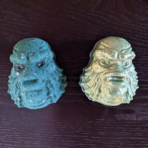 May include: Two bath bombs shaped like the Creature from the Black Lagoon. One is green and the other is gold.