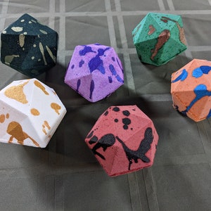 May include: Five geometric polyhedral bath bombs in various colors including black with gold, purple with dark blue, green with copper, white with gold, orange with blue, and red with black paint splatter patterns arranged on a dark surface.