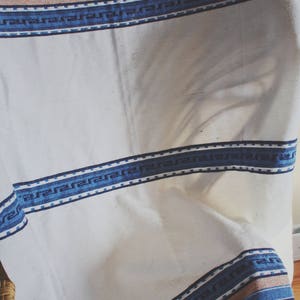 May include: White fabric tablecloth with a blue and white patterned border. The tablecloth is draped over a chair.