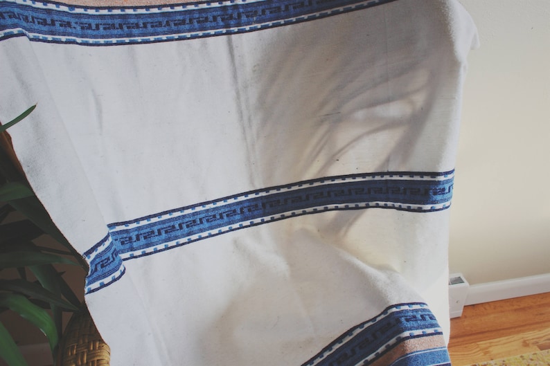 May include: White fabric tablecloth with a blue and white patterned border. The tablecloth is draped over a chair.