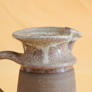 May include: A brown and white ceramic pitcher with a textured glaze. The pitcher has a wide, round spout and a handle.